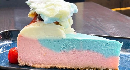 A slice of colorful cake with blue, pink, and white layers, topped with cotton candy and a cherry on a dark plate.