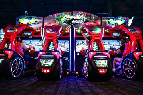 Two racing arcade machines with vibrant lights and screens, featuring players engaged in a racing game.