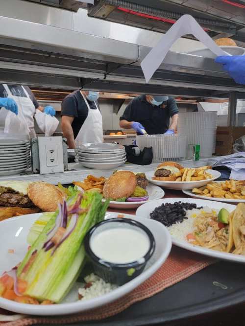 A busy kitchen scene with plates of food in the foreground and chefs preparing meals in the background.