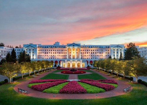 A grand hotel with a colorful garden at sunset, featuring blooming flowers and a beautifully lit facade.