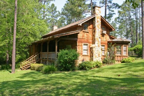 A rustic wooden cabin surrounded by trees and greenery, featuring a stone chimney and a welcoming porch.