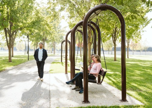 A man stands nearby while two people sit on a swing set in a sunny park surrounded by trees.