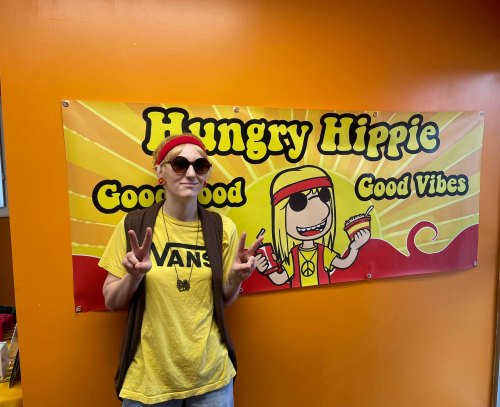 A person in sunglasses and a yellow shirt poses peace sign in front of a colorful "Hungry Hippie" banner.