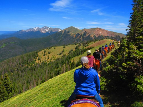 A group of horseback riders on a scenic trail, surrounded by lush green hills and distant snow-capped mountains.