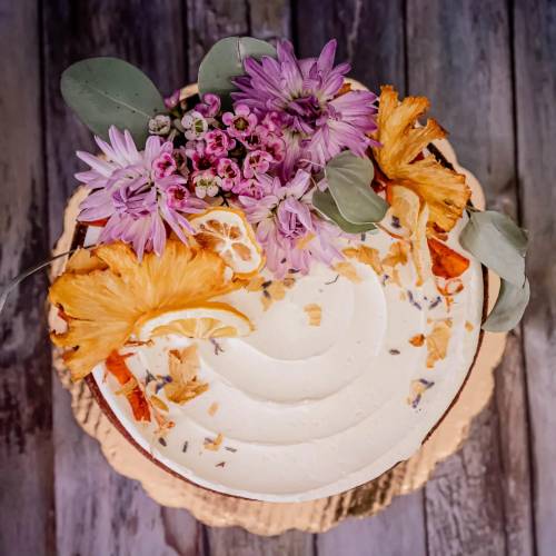 A beautifully decorated cake topped with flowers, dried orange slices, and coconut flakes on a wooden surface.