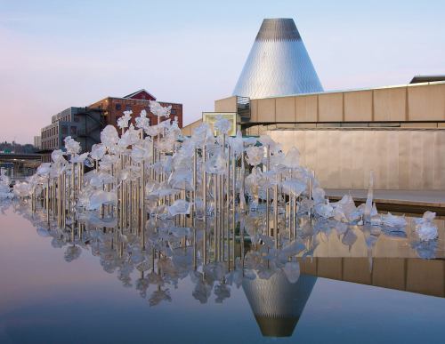 A modern building with a cone-shaped roof reflects in water, surrounded by artistic glass sculptures.
