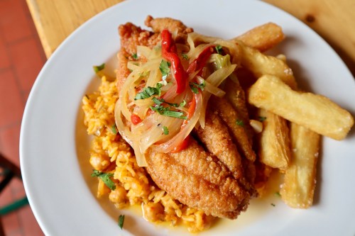 A plate of fried fish topped with sautéed onions and peppers, served with rice and fried plantains.