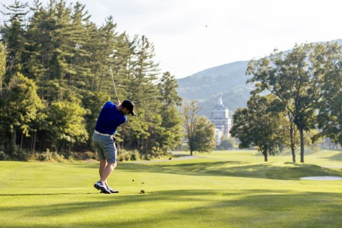 A golfer swings on a lush green course with trees and mountains in the background, capturing a sunny day.