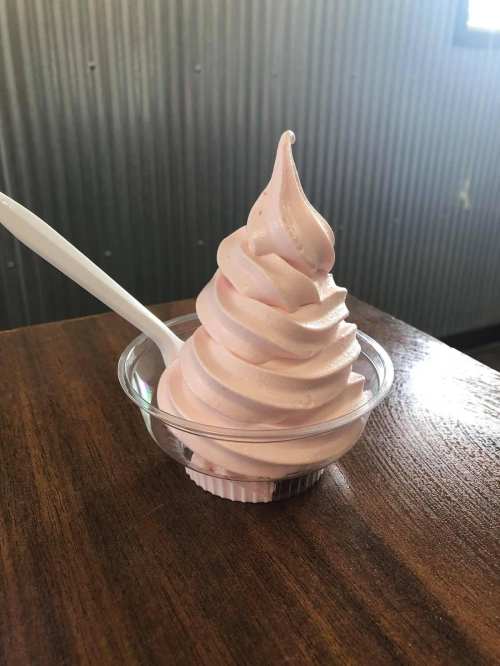 A swirl of pink soft serve ice cream in a clear cup, resting on a wooden table with a plastic spoon.