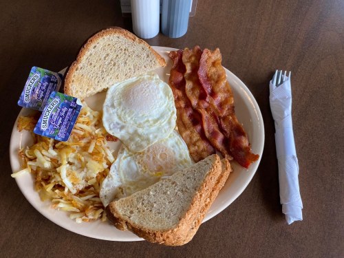 A plate of breakfast featuring two fried eggs, crispy bacon, hash browns, and two slices of toast with condiments.