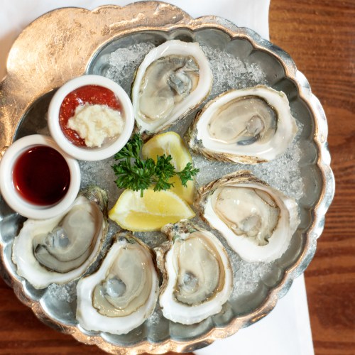 A silver platter with six fresh oysters on ice, accompanied by lemon wedges, cocktail sauce, and horseradish.