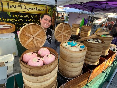 A vendor smiles behind bamboo steamers filled with colorful bao buns at a market stall.