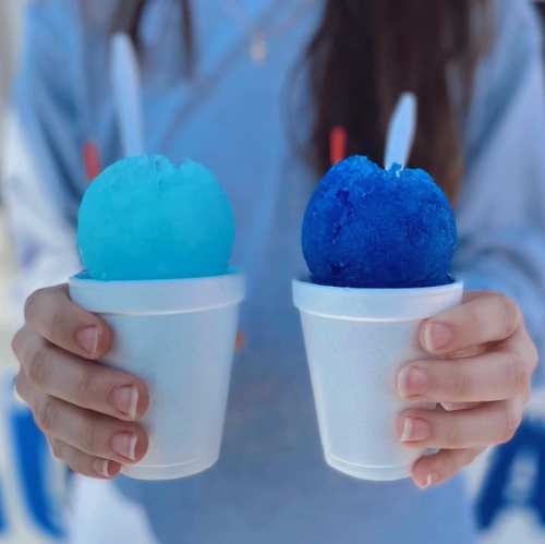 A person holds two cups of blue snow cones, one light blue and one dark blue, with spoons in each cup.