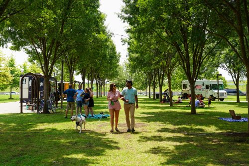 A couple walks a dog through a park with food trucks and people enjoying the outdoors under green trees.