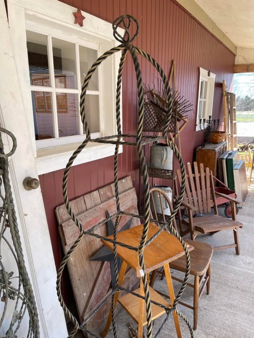 A porch filled with vintage furniture and decor, including a twisted metal stand, wooden chairs, and various antiques.