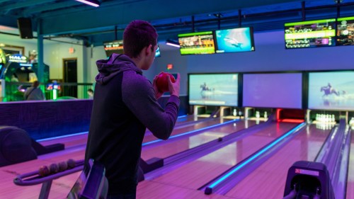 A person prepares to bowl in a modern bowling alley with colorful lighting and screens displaying scores.