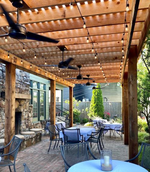 Outdoor patio with string lights, wooden beams, and tables set for dining, featuring a stone fireplace and greenery.