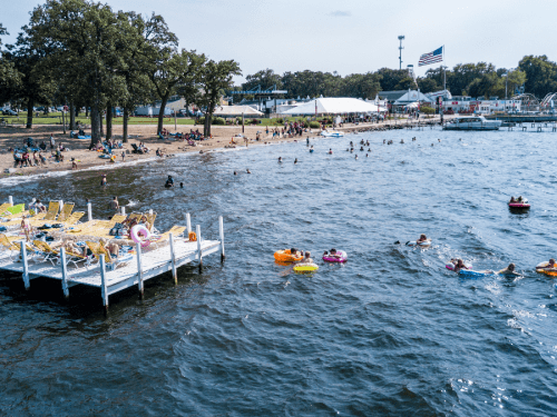 A busy beach scene with people swimming, lounging on floats, and enjoying the sun near a dock and amusement area.
