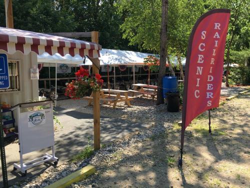 A scenic outdoor area with a tent, picnic tables, and a flag for Scenic Railriders, surrounded by greenery.