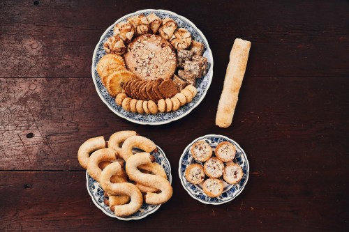 A variety of baked goods on decorative plates, including cookies, pastries, and a long breadstick, on a wooden table.