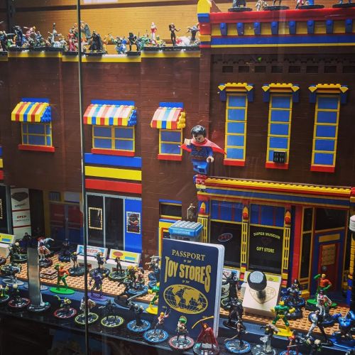A colorful display of toy figures and buildings, featuring superheroes and a toy store theme in a glass case.