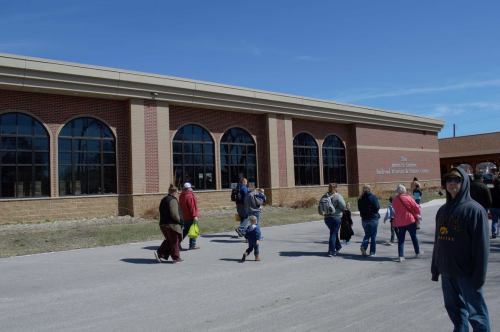 A group of people walking outside a large brick building with arched windows on a sunny day.