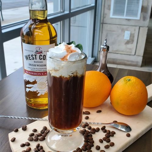 A glass of coffee topped with whipped cream, beside a bottle of whiskey and fresh oranges on a wooden board.