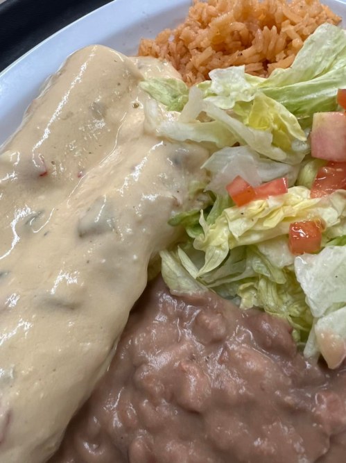 A plate of Mexican food featuring enchiladas with creamy sauce, rice, refried beans, and a side salad with tomatoes.