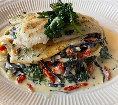 A plate of grilled fish atop black pasta with spinach, sun-dried tomatoes, and a creamy sauce, garnished with herbs.
