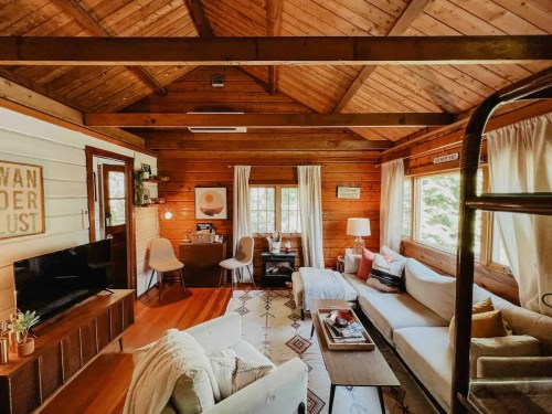 Cozy wooden cabin interior with a living area, desk, and large windows, featuring warm lighting and rustic decor.