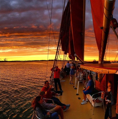 Sailing boat at sunset with passengers enjoying the view and vibrant orange and purple skies reflected on the water.