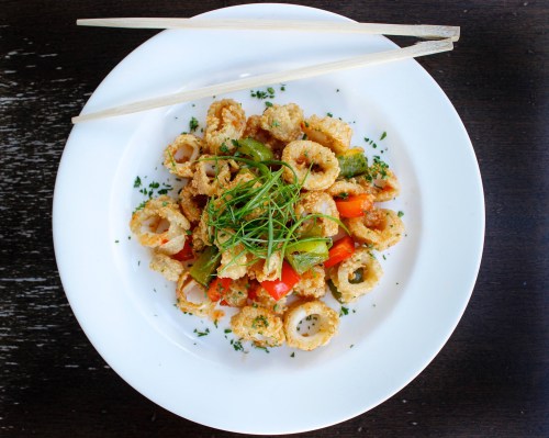 A plate of crispy fried calamari with bell peppers and green onions, garnished with herbs, served with chopsticks.