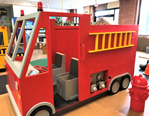 A colorful red toy fire truck with two seats, a fire hydrant, and decorative lights, set in a play area.