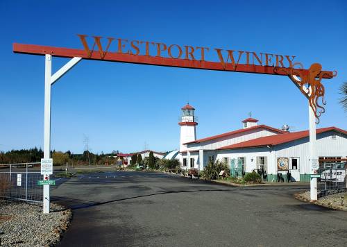 Sign for Westport Winery with a lighthouse and winery buildings in the background under a clear blue sky.