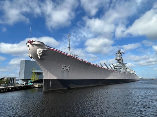 A large battleship, numbered 64, docked in calm waters under a partly cloudy sky.