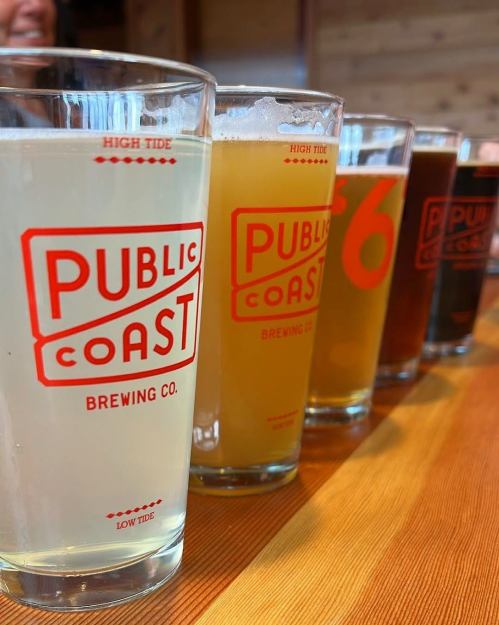 Five glasses of beer with the logo "PUBLIC COAST Brewing Co." on a wooden table.