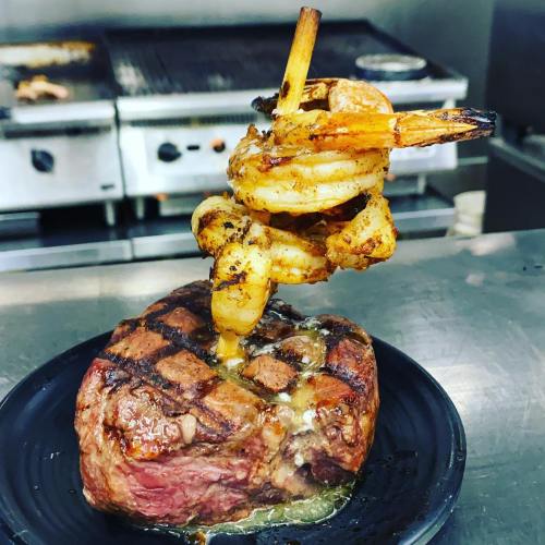 A grilled steak topped with shrimp skewers, served on a black plate in a kitchen setting.