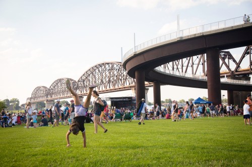 A child performs a handstand on grass at a lively outdoor event near a bridge and a crowd of people.