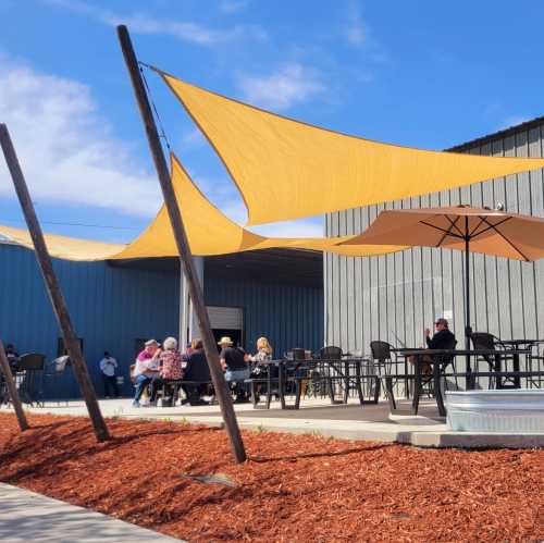 A sunny outdoor seating area with yellow shade sails, people dining, and a modern building in the background.