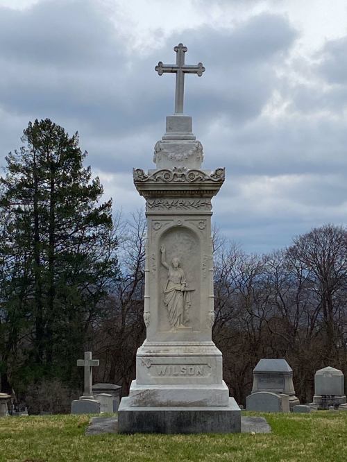 A tall, ornate gravestone with a cross on top, featuring a carved figure and the name "WILSON" at the base.