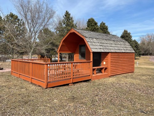 A small orange cabin with a sloped roof and wooden deck, surrounded by trees and grass under a clear blue sky.