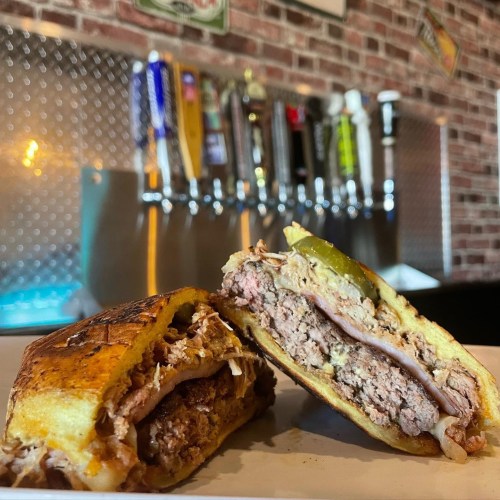 A close-up of a sliced sandwich filled with meats and pickles, served on a plate with a bar backdrop.