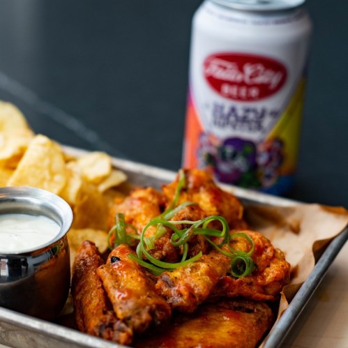 A tray of spicy chicken wings with green onions, a side of ranch dressing, potato chips, and a colorful beverage can.