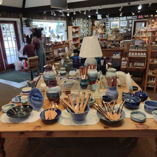 A cozy shop interior featuring a wooden table with various colorful pottery and kitchenware, surrounded by shelves of goods.