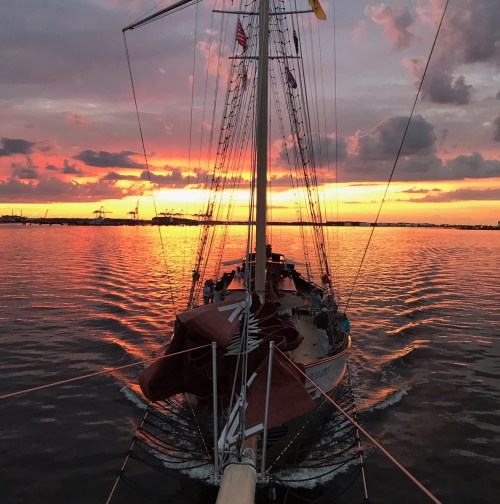 A sailboat on calm waters at sunset, with vibrant orange and purple skies reflecting on the surface.
