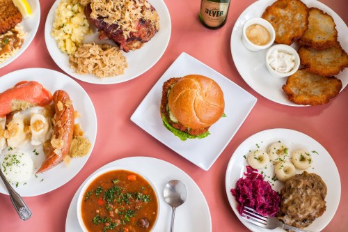 A colorful spread of various dishes including sandwiches, salads, and sides on white plates against a pink tablecloth.