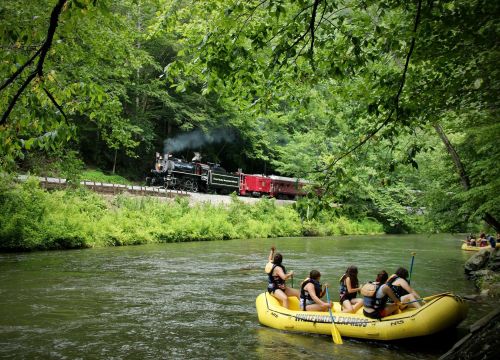 A steam train travels alongside a river where people in yellow rafts enjoy a summer day in a lush green setting.