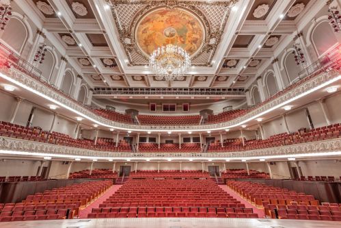 Interior of a grand theater featuring ornate architecture, a chandelier, and red seating arranged in tiers.