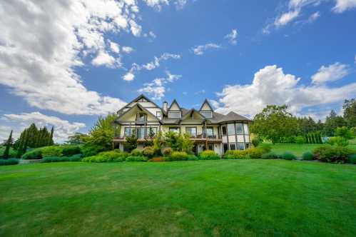 A large, elegant house surrounded by lush green lawns and trees under a bright blue sky with fluffy clouds.