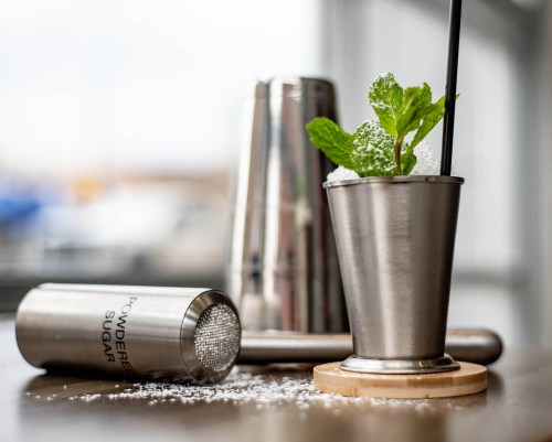 A metal cocktail shaker and a mint julep cup with mint leaves, set on a wooden coaster with powdered sugar scattered around.